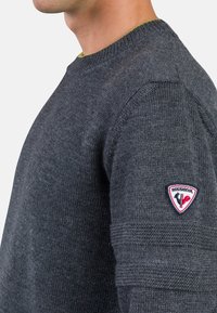 Dunkelgrauer Strickpullover mit Rundhalsausschnitt, gerippten Bündchen und einem kleinen gestickten Logo am linken Ärmel. Strukturierte Stoffdetails sind sichtbar.