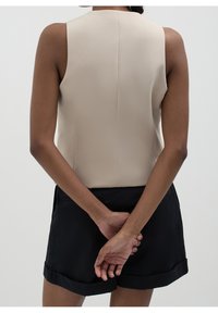 Personne vue de dos portant un haut beige sans manches et un short noir, les mains jointes dans le dos.
