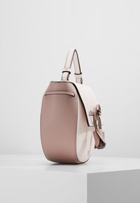 Petit sac à main bicolore en cuir beige et marron clair avec poignée supérieure, rabat avant et nœud décoratif en cuir, posé sur un socle blanc.