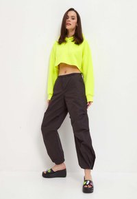 Hanorac scurt, neon galben, cu mâneci lungi, asortat cu pantaloni negri largi. Sandalele negre au un design cu toc și accente colorate.
