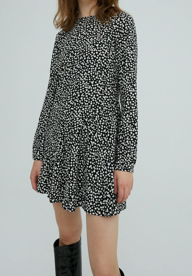 Svart långärmad klänning med ett vitt blommönster, som har rund halsringning och utsvängd nederkant. Modellen har matchat den med knähöga svarta stövlar.