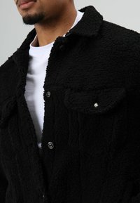 Zwarte nepbontjas met een zachte, gestructureerde afwerking, voorzien van zilveren drukknopen en twee borstzakken. Draagt over een wit T-shirt.