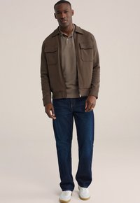 Bruine jas met rits en twee zakken aan de voorkant, daaronder een taupekleurig poloshirt, een donkerblauwe spijkerbroek en witte sneakers met beige zolen.