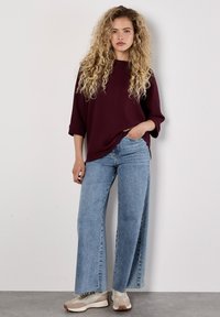 Bordeaux waffle-knit oversized trui met driekwartmouwen, gecombineerd met lichtblauwe high-waisted wijde jeans en beige sneakers.