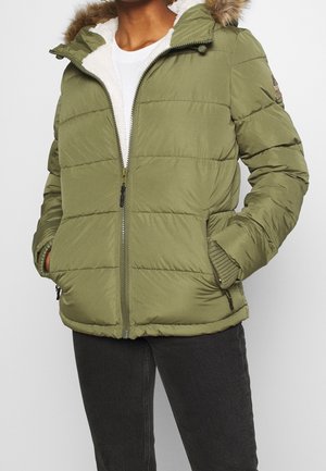 Personne portant une doudoune vert olive avec une capuche bordée de fourrure, une fermeture éclair à l'avant, et les mains dans les poches d'un pantalon noir.