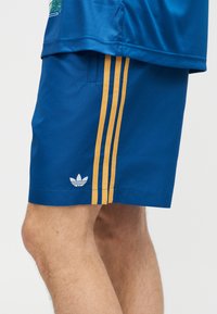 Μπλε αθλητικό σορτς με τρεις κάθετες κίτρινες ρίγες και λευκό λογότυπο Adidas κοντά στο τελείωμα στο πόδι ενός ατόμου.