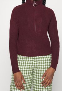 Persona vistiendo un suéter de canalé burdeos con cremallera y tirador en forma de anillo, combinado con pantalones de patrón de pata de gallo en verde y blanco.