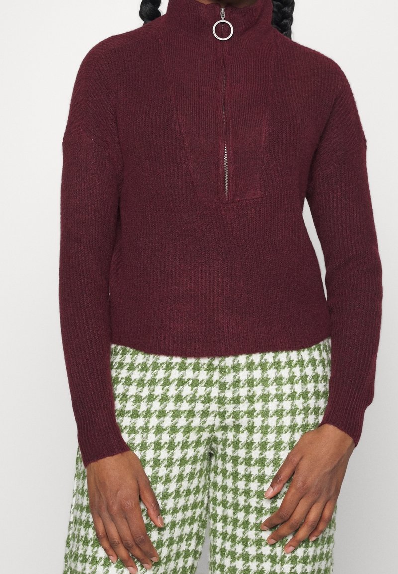 Persona vistiendo un suéter de canalé burdeos con cremallera y tirador en forma de anillo, combinado con pantalones de patrón de pata de gallo en verde y blanco.