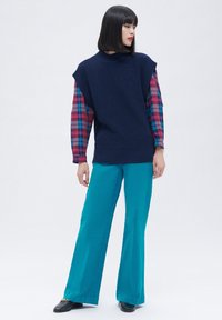 Laivaston sininen neule sleeveless-toppi, joka on puettu päälle ruudullisen pitkähihaisen paidan kanssa, yhdistettynä turkoosisiin leveälahkeisiin housuihin ja mustiin kengiin, joissa on ketjuakoriste.