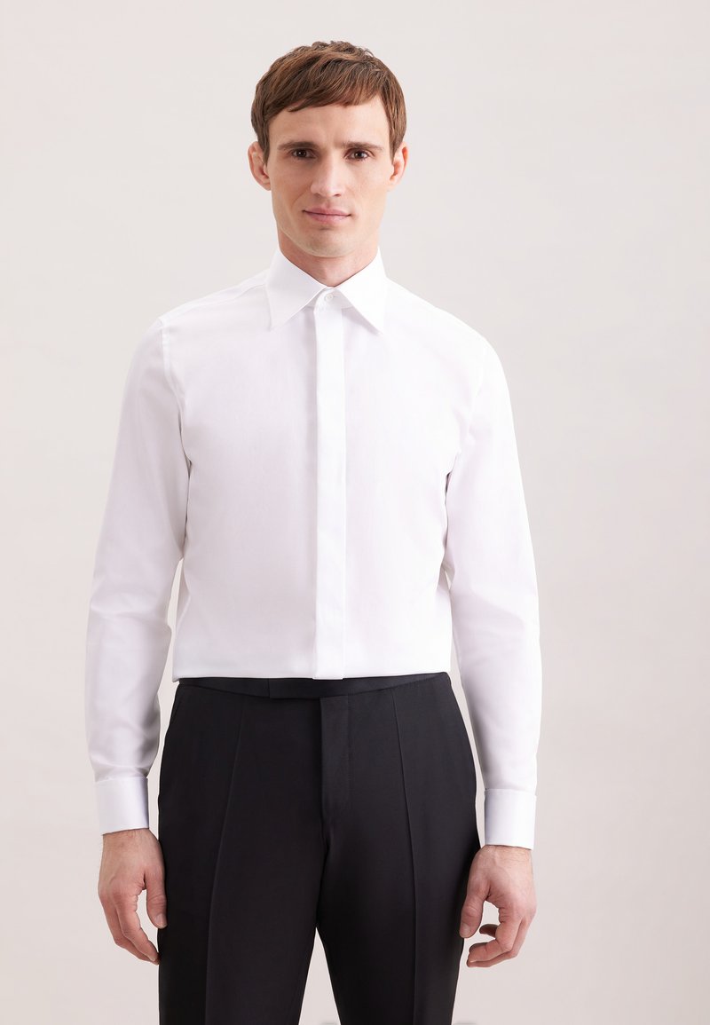 Homme portant une chemise blanche cintrée avec des boutons cachés et un pantalon de costume noir, debout devant un fond clair uni.