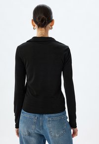 Schwarzes Langarmshirt mit Kragen, aus einem glatten Material gefertigt. Kombiniert mit blauen Jeans aus Denim, die Hintertaschen und eine abgenutzte Textur aufweisen.