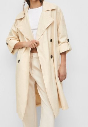Person trägt einen beigen Trenchcoat mit schwarzen Knöpfen über einem weißen Hemd und beigen Hosen und hält mit einer Hand das Revers des Mantels.