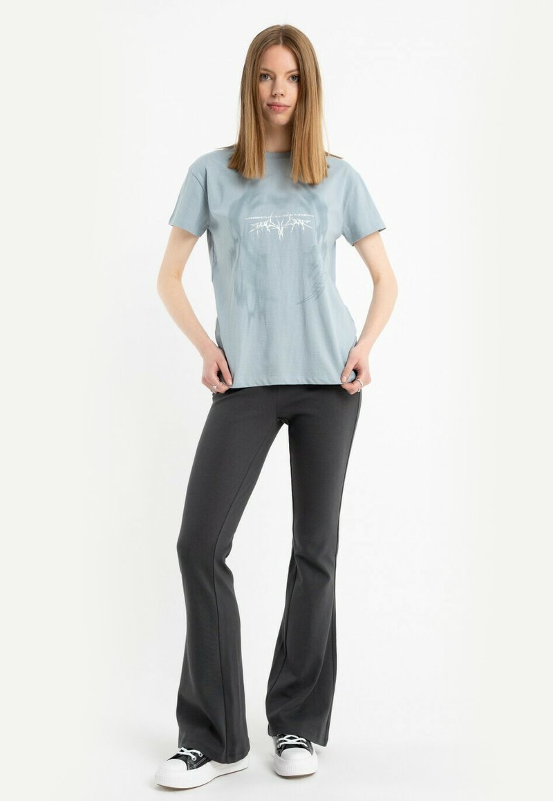 Kobieta z prostymi, jasnobrązowymi włosami, ubrana w jasnoniebieską koszulkę z nadrukiem, czarne rozszerzane spodnie i czarno-białe trampki, stoi z rękami na biodrach.