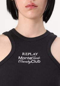 Zwarte geribbelde tanktop met een ronde halslijn en mouwloos ontwerp, versierd met witte tekst: "REPLAY MonteCarlo CountryClub."