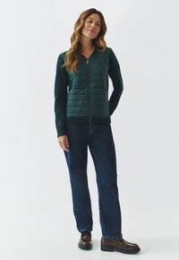 Green zip-up jacket with padded front, ribbed sleeves, and dark blue straight-leg jeans. Model wears brown lace-up boots.