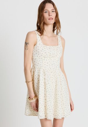 Giovane donna che indossa un vestito crema senza maniche con una piccola stampa floreale e scollo quadrato, con braccialetti sul braccio sinistro e un tatuaggio a fiocco sul braccio destro.