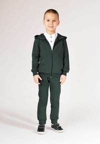 Zielona bluza z kapturem i dopasowane spodnie dresowe wykonane z miękkiego materiału. Bluza posiada przedni zamek błyskawiczny i dwie boczne kieszenie. Buty sportowe z czarnymi akcentami.