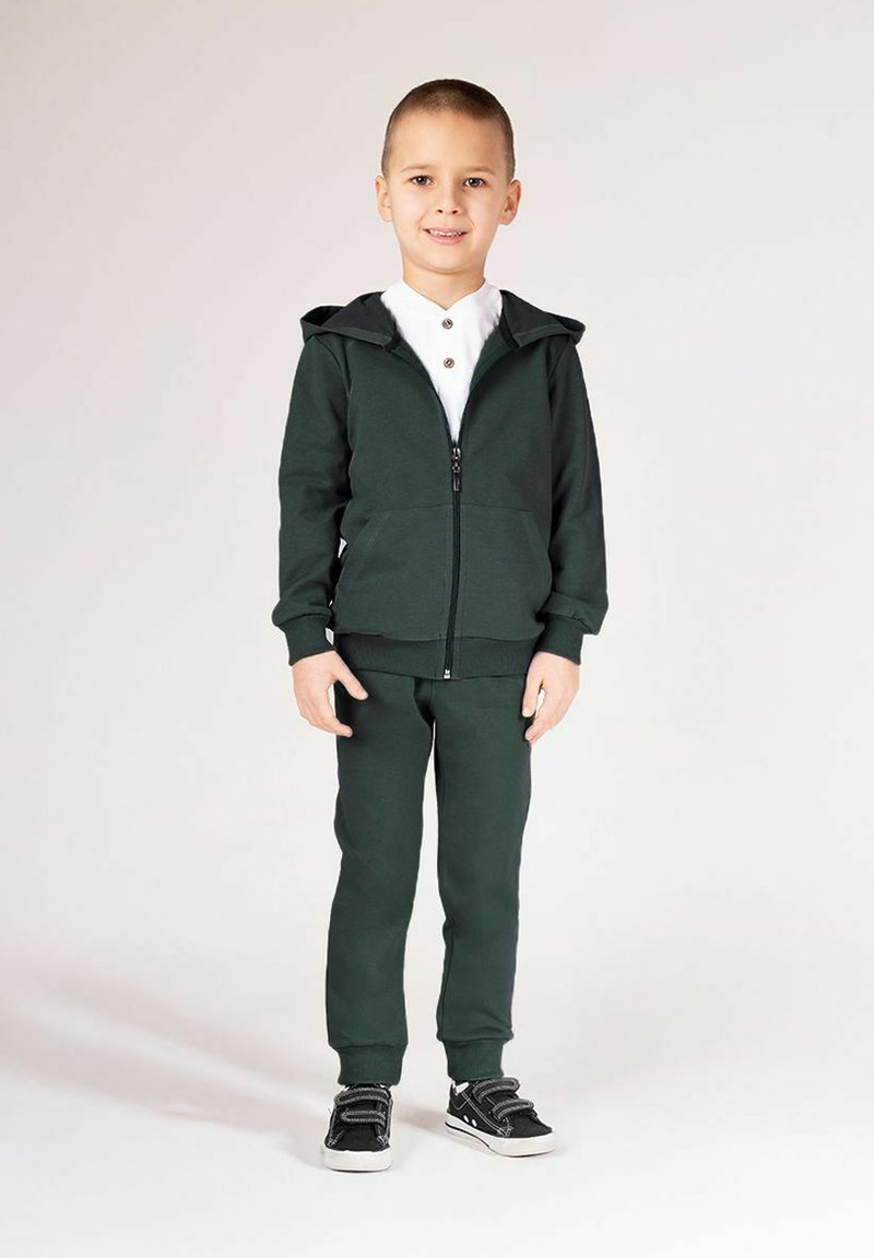 Zielona bluza z kapturem i dopasowane spodnie dresowe wykonane z miękkiego materiału. Bluza posiada przedni zamek błyskawiczny i dwie boczne kieszenie. Buty sportowe z czarnymi akcentami.