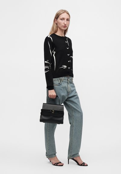 Femme debout portant un haut manches longues noir à motifs, un jean bleu clair, des sandales noires à talons et tenant un sac à main noir.
