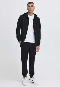 Sort hættejakke med lynlås foran og snore, parret med matchende sorte joggers. Hvid t-shirt under og hvide sneakers med sorte striber.