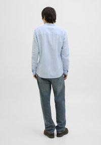 Camisa de manga larga azul clara con detalle en el hombro trasero, puños abotonados y dobladillo curvado, combinada con jeans azul claros y zapatos marrones.