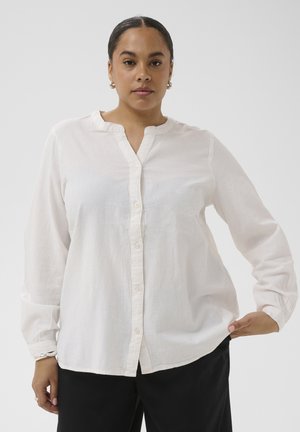 Vrouw draagt een witte blouse met knopen en lange mouwen en zwarte broek, staand tegen een effen lichte achtergrond.