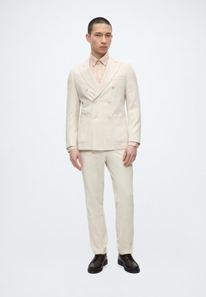 Muž oblečený v krémovom dvojradovom obleku s bledoružovou košeľou a tmavohnedými topánkami, stojaci pred jednoduchým bielym pozadím.