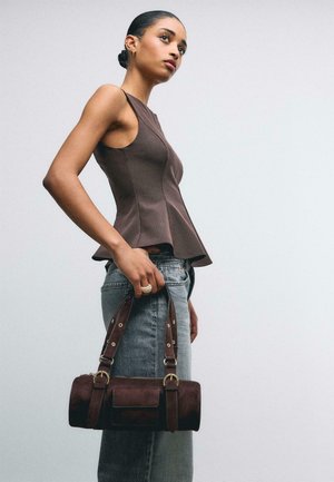 Femme portant un haut marron sans manches et un jean taille haute, tenant un sac à main cylindrique marron foncé sur un fond clair uni.