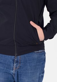 Giacca nera con zipper, dalla texture liscia, polsini elasticizzati e un discreto dettaglio del logo. Tasca frontale visibile sul lato sinistro.