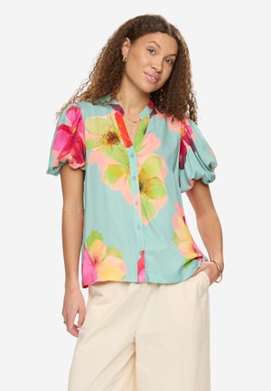 Kobieta o kręconych włosach, ubrana w turkusową bluzkę z dużymi kolorowymi kwiatowymi wzorami oraz beżowe spodnie o szerokich nogawkach, stojąca z jedną ręką w kieszeni.