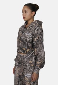 Hoodie court dans un motif camouflage avec des nuances de brun et de gris, doté d'une poche avant et d'un détail de logo. Associé à un pantalon camouflage assorti.