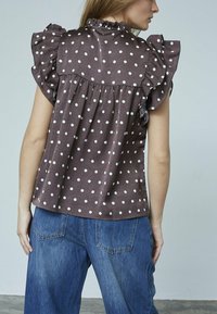 En kvinde står med ryggen til kameraet, iført en brun bluse med prikkede mønstre og volanske ærmer samt blå jeans, mod en neutral baggrund.