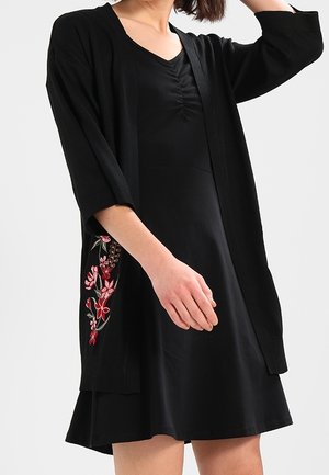 Femme portant une robe noire avec un col froncé et un cardigan noir orné de broderies florales rouges et blanches sur le côté.
