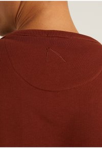 Primo piano della schiena di una persona che indossa una maglietta color ruggine con colletto a coste e un piccolo dettaglio cucito curvo sotto il décolleté.