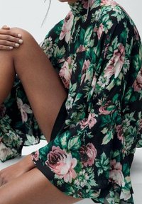 Robe noire à motifs floraux avec col haut, manches larges et un design fluide. Elle arbore de grands motifs de fleurs roses et vertes sur un tissu transparent.