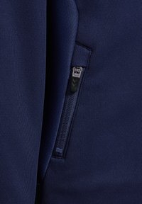 Marinblått tyg med en slät textur har en dold zipficka med en svart dragkedja. Sömdetaljer längs kanterna förbättrar designen.