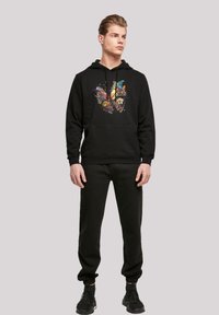 Svart huvtröja med en färgglad fjäril och blommönster på framsidan, i kombination med svarta joggers. Materialet verkar mjukt och texturerat.