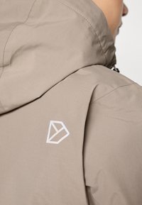 Beež veekindel jope, millel on kapuuts, sile tekstuur ja valge geomeetriline logo seljal. Minimalistlik disain, ilma nähtava riistvarata.