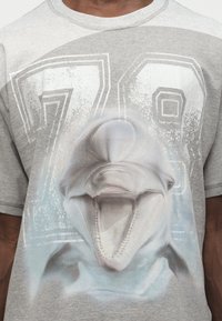 Κοντινό πλάνο ενός γκρι T-shirt με μεγάλο γραφικό μιας κεφαλής δελφινιού και τον ξεθωριασμένο αριθμό 78 στο παρασκήνιο.