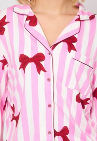 Camisa de pijama a rayas pink y blanca con estampado de lazo rojo, cierre de botones, cuello con muesca y bolsillo. Presenta un ribete de contraste a lo largo de los bordes.