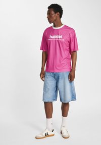Magenta kortärmad t-shirt med vit krage och texten "hummel PERFORMANCE", tillsammans med ljusblå denimshorts och vita sneakers.