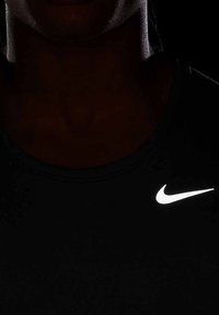 Personne portant un t-shirt noir avec un logo blanc Nike swoosh proéminent sur la poitrine, dans une lumière faible où seul le logo est clairement visible.