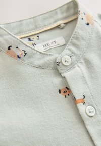 Camisa de tela verde claro con cuello abotonado, con patrones de perros de dibujos animados en naranja y negro. La etiqueta dice "NEXT".