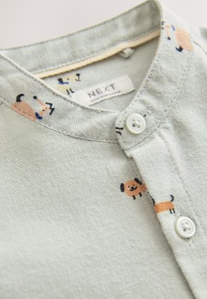 Svetlozelená látková košeľa s golierom na gombíky, zdobená karikatúrnymi vzormi psov v oranžovej a čiernej farbe. Štítok nesie nápis "NEXT".