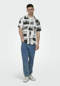 Camicia a maniche corte con bottoni in bianco e nero con stampa fotografica; abbinata a jeans in denim blu e sneakers bianche.
