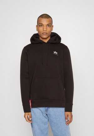 Schwarzer Baumwollhoodie mit einer Kängurutasche, Kapuze mit Kordelzug und einem kleinen weißen Logo auf der Brust. Kombiniert mit hellblauen Jeans.
