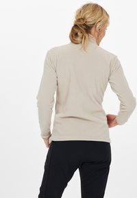 Donna con capelli biondi raccolti in un bun basso e disordinato, che indossa una giacca in pile beige e pantaloni neri, in piedi di spalle con una mano sui fianchi.