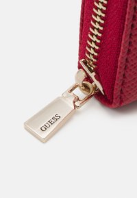 Rode gestructureerde ritstas met gouden hardware en een zilveren label met de tekst "GUESS." Bevat een opvallende gouden rits en gedetailleerde stiksels.