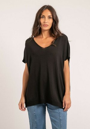 Femme portant un haut ample noir à manches courtes et col en V, et un jean bleu, debout devant un fond clair uni.