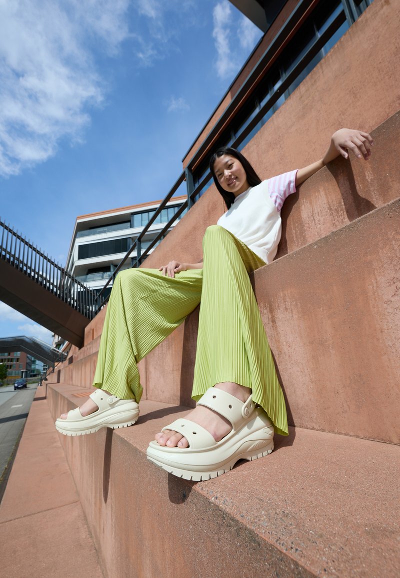 Light beige sandals with dual straps, a wedge sole, and a textured upper. The individual wears pleated, vibrant green pants and a white top.